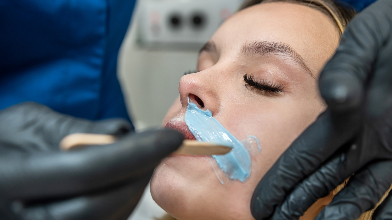 Woman receiving upper lip waxing, blue wax applied, gloved hands, close-up.