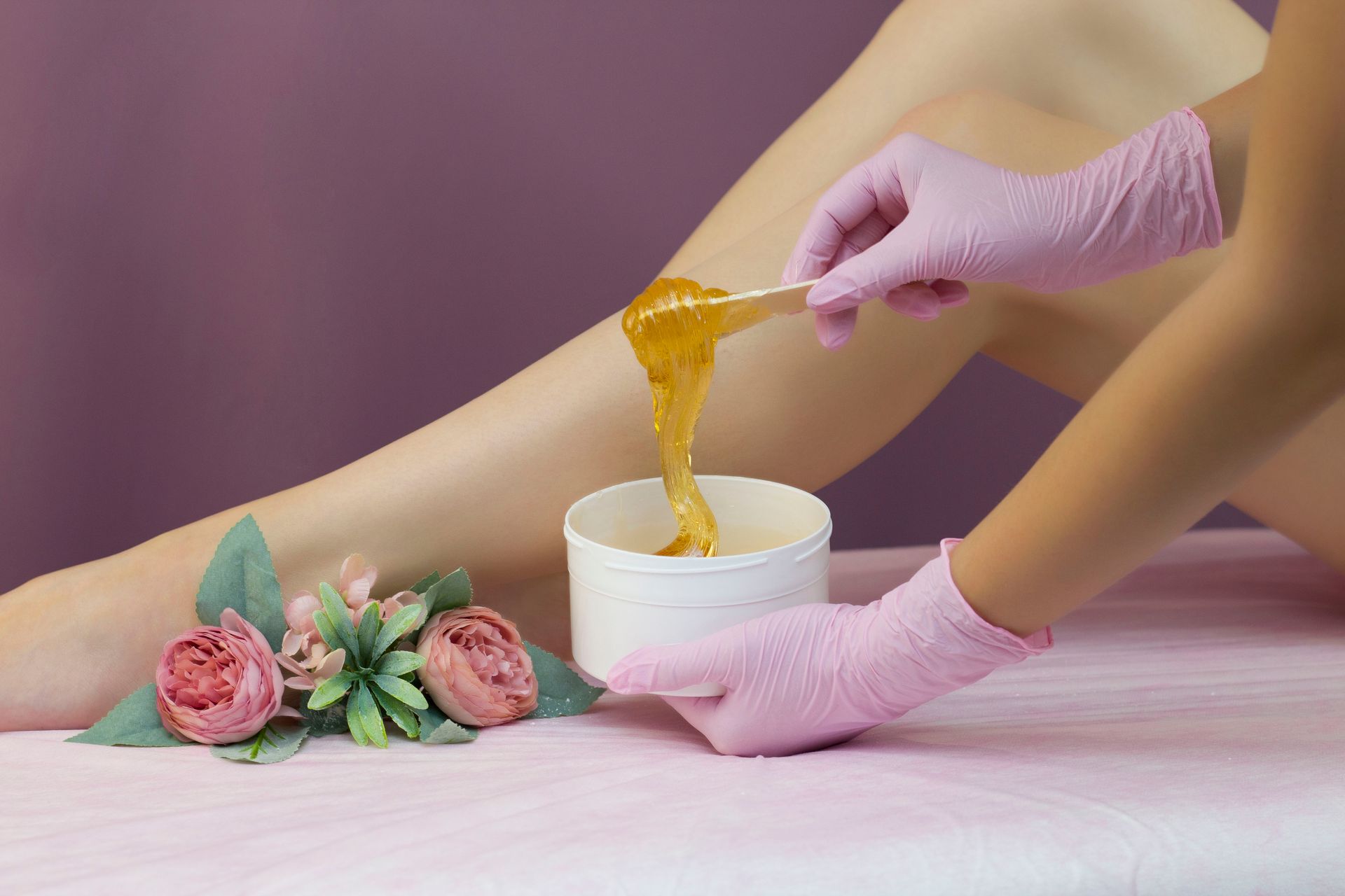 Person applying wax to leg with spatula. White bowl, pink gloves, leg, pink flowers, and table are visible.
