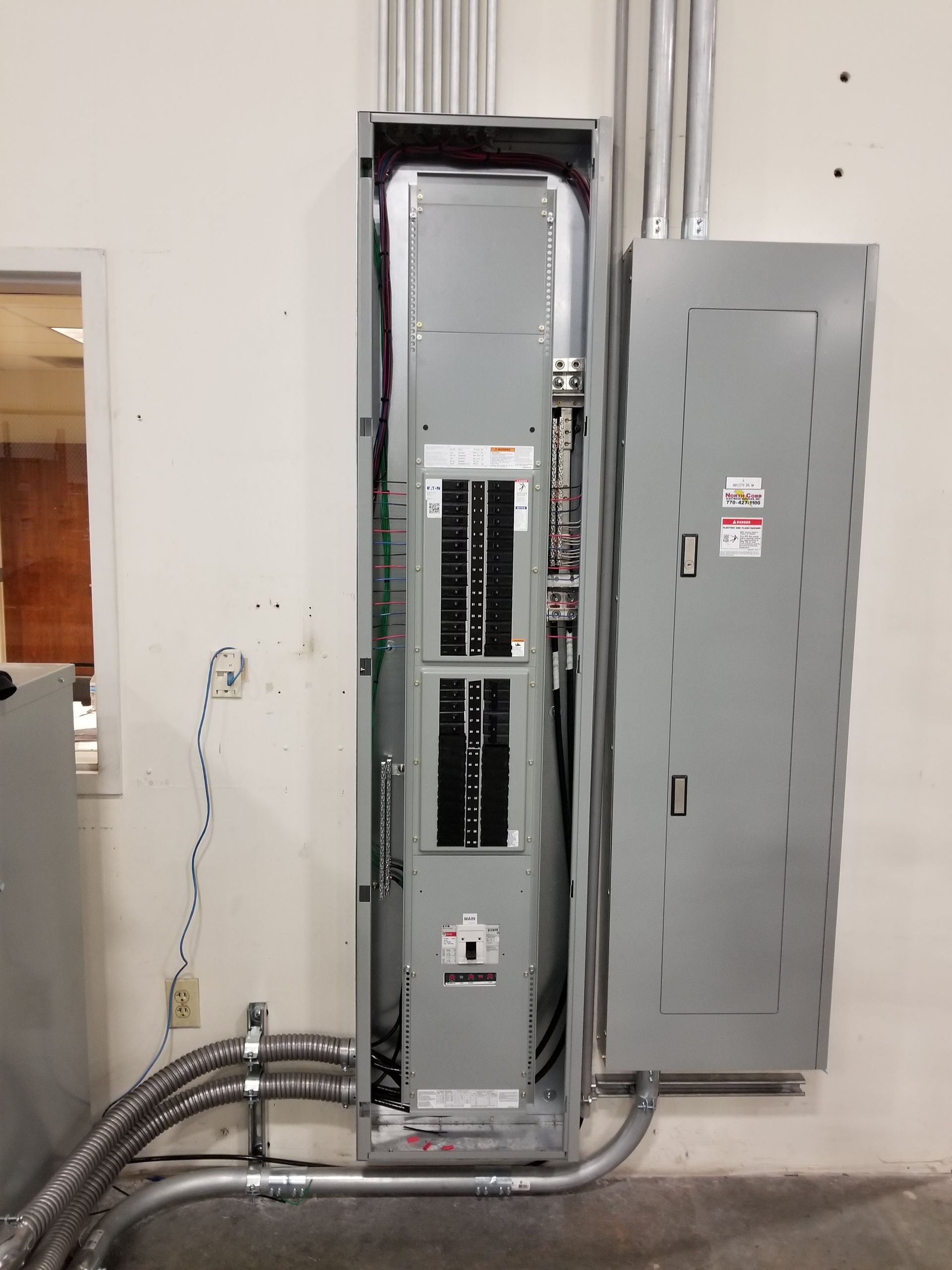 Electrical panel with open door, mounted on a white wall. Gray conduits and wires are visible.