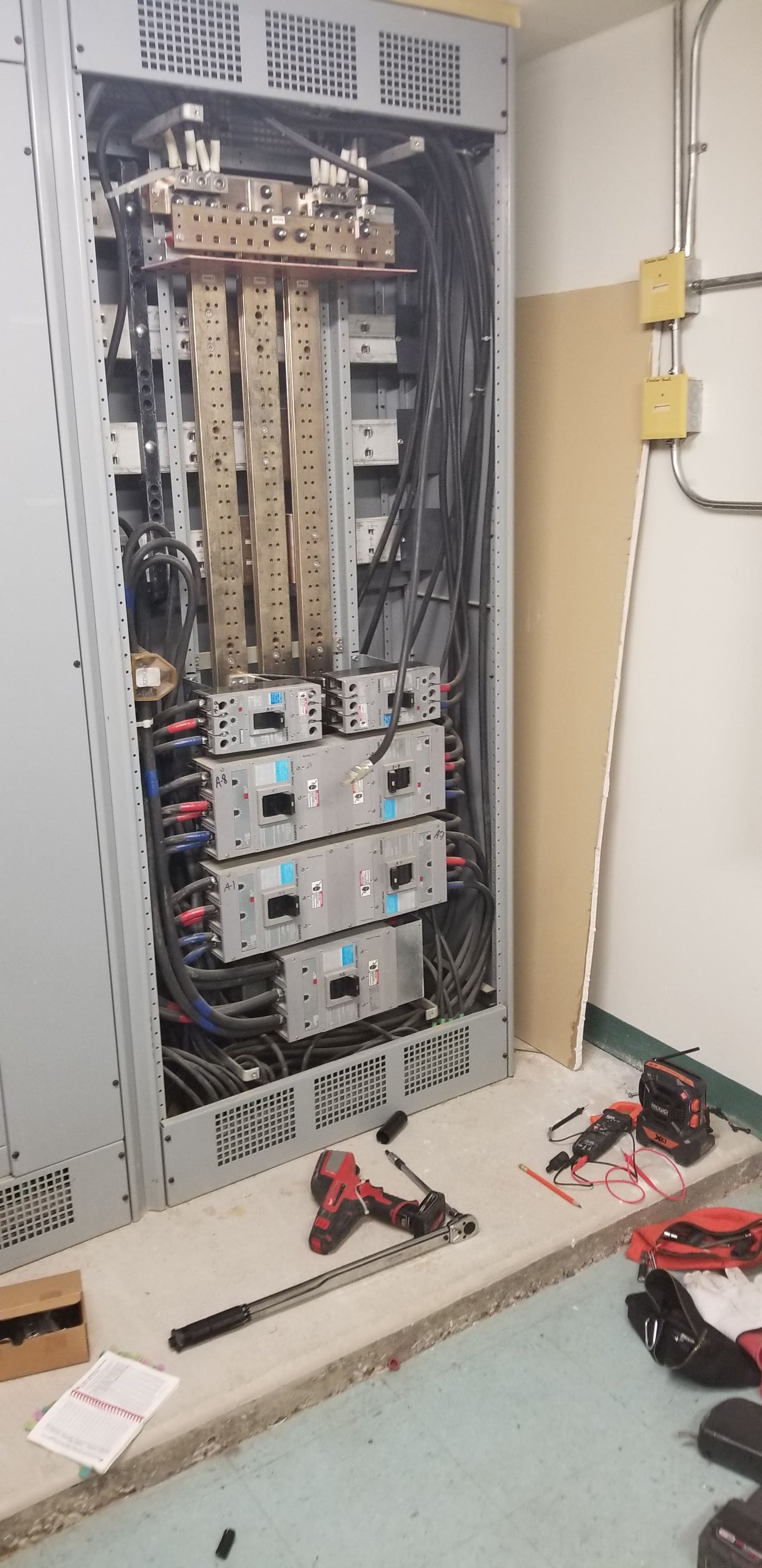 Open electrical panel with wires and circuit breakers; tools on floor.