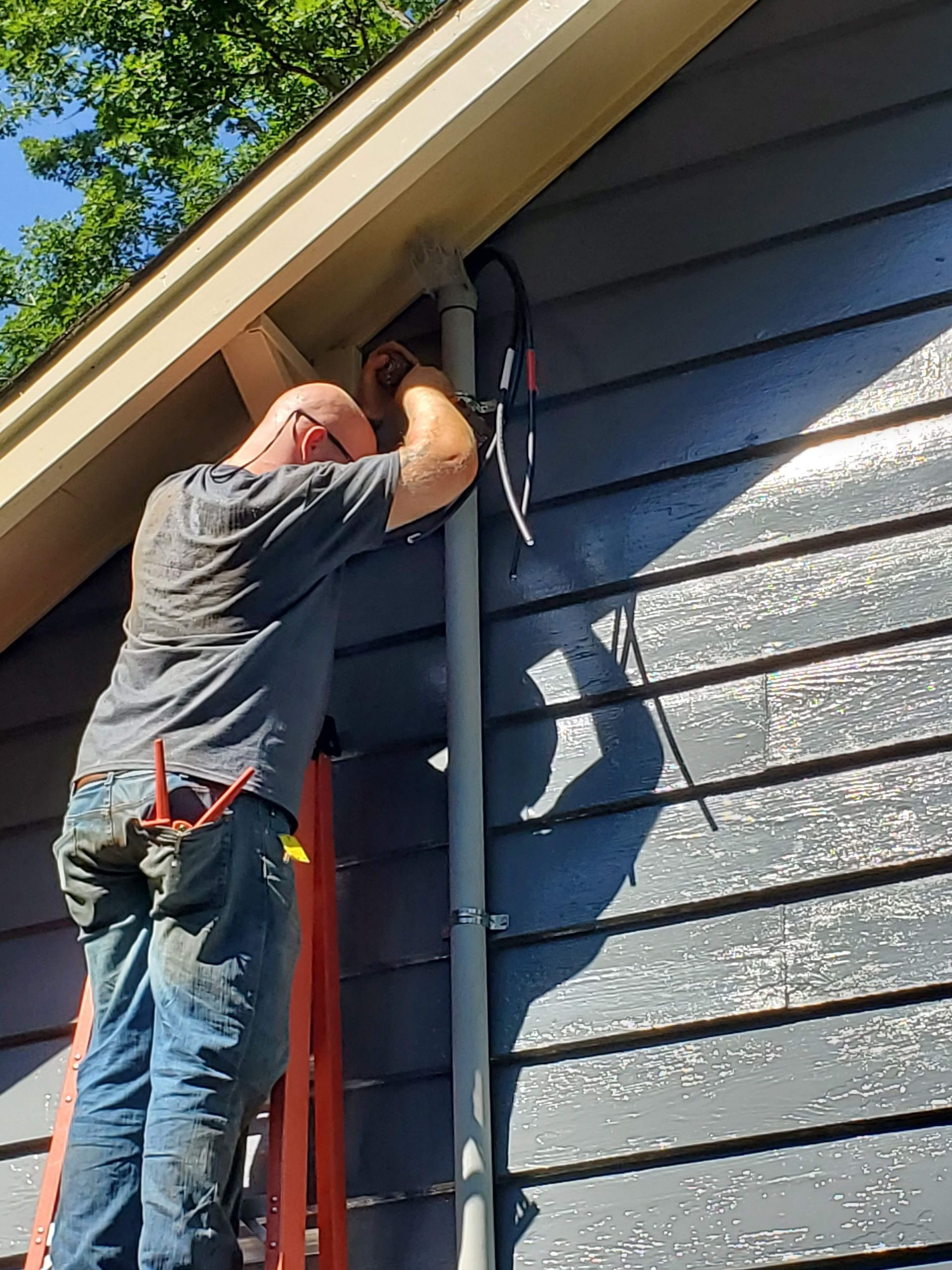 Man on a ladder installing cables on a gray-sided building with white trim.