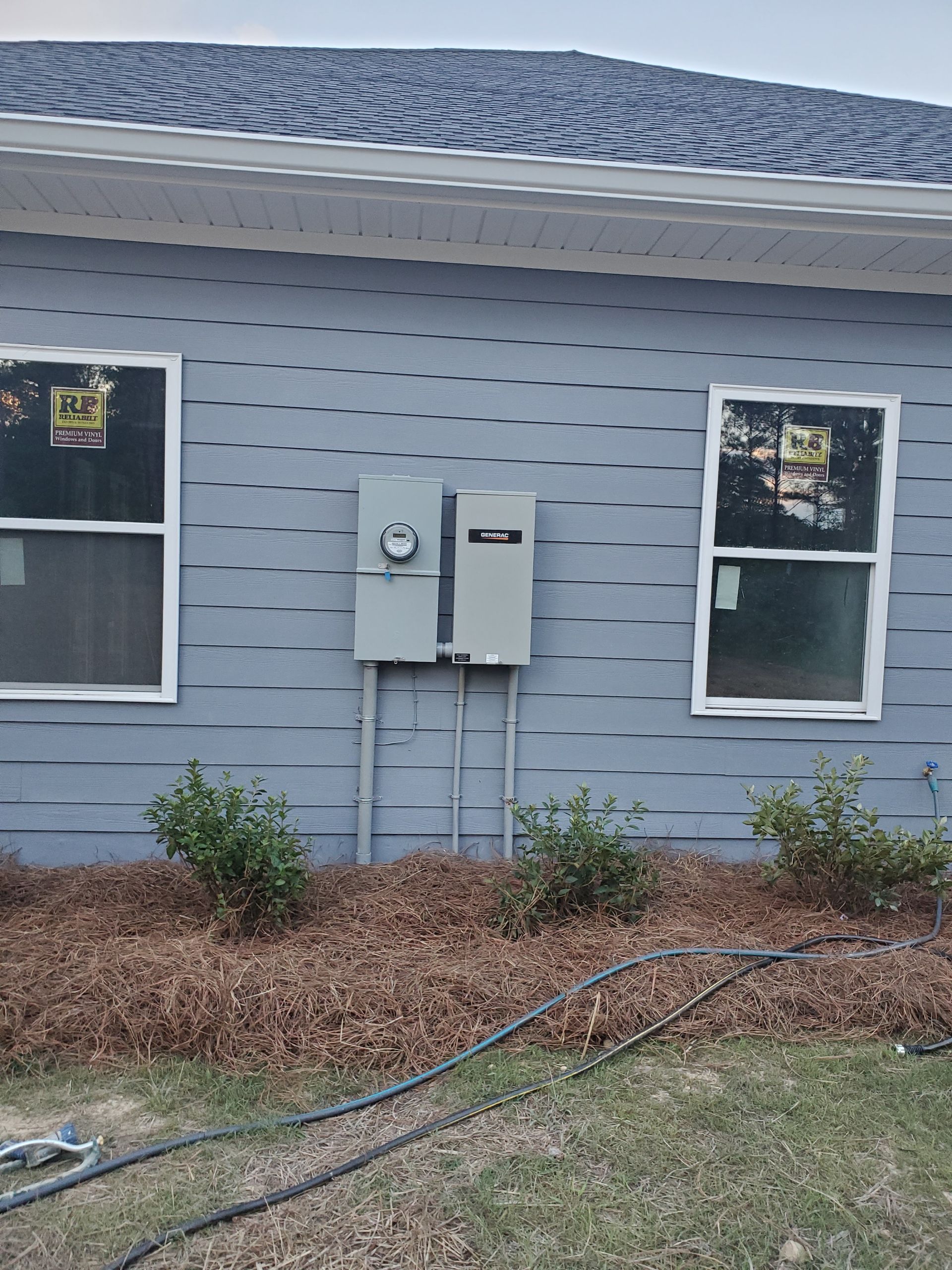 Gray house exterior with electrical boxes, windows, and mulch.