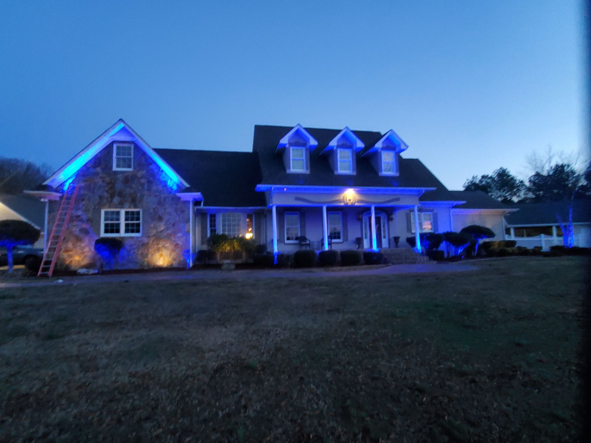 A house with blue lights illuminating the roof, trim, and landscaping at dusk.