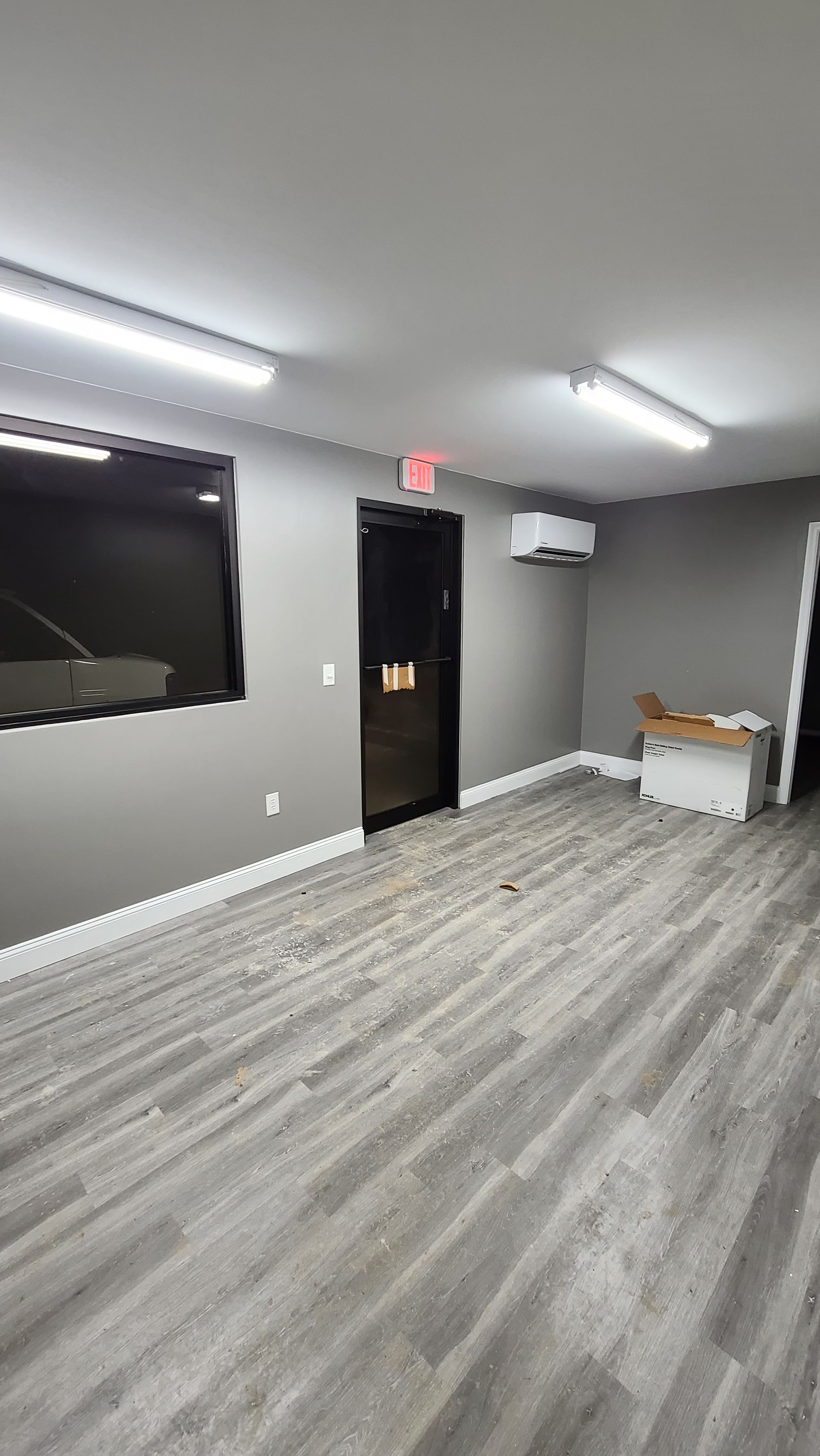 Empty room with grey walls and wood-look flooring. A TV, door, and air conditioning unit are visible.