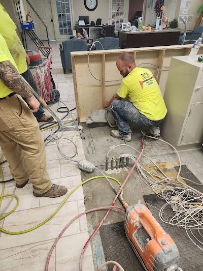 Men in yellow shirts cutting flooring near a wood structure with equipment and wires in a room.