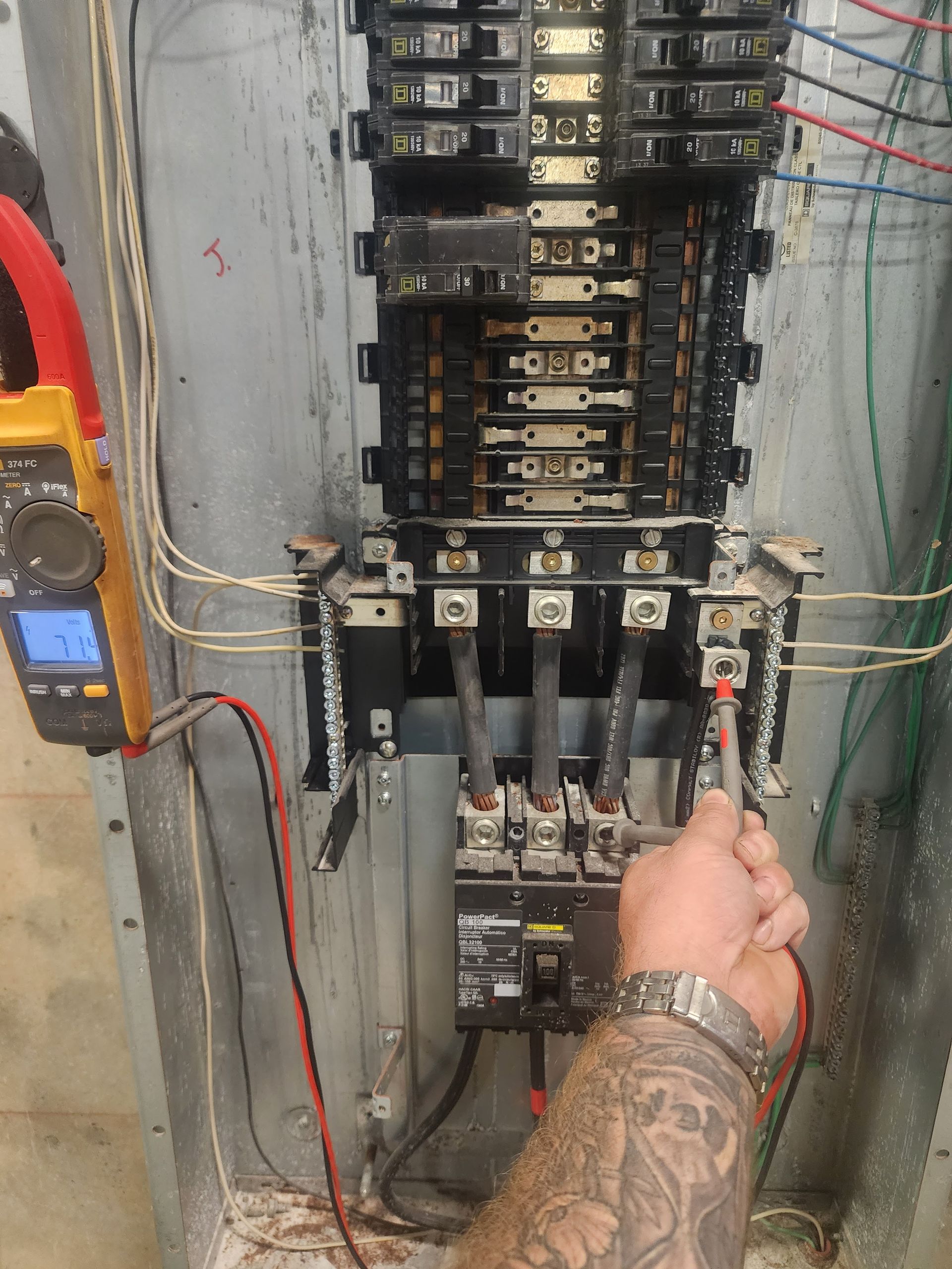 Electrician testing voltage at a breaker panel with a multimeter. Grey panel, black wires, hand visible.