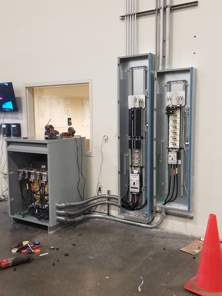 Electrical panel installation with exposed wires, conduit, and orange safety cone in a construction setting.