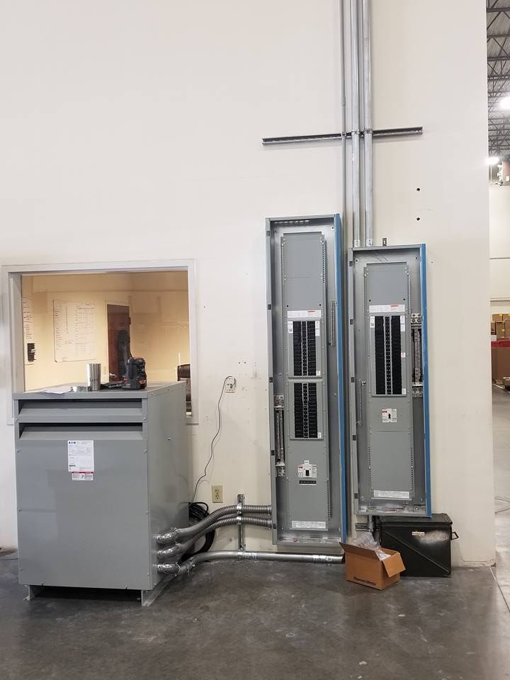 Electrical equipment installed on a white wall in a warehouse; includes transformer, electrical panels, and conduit.