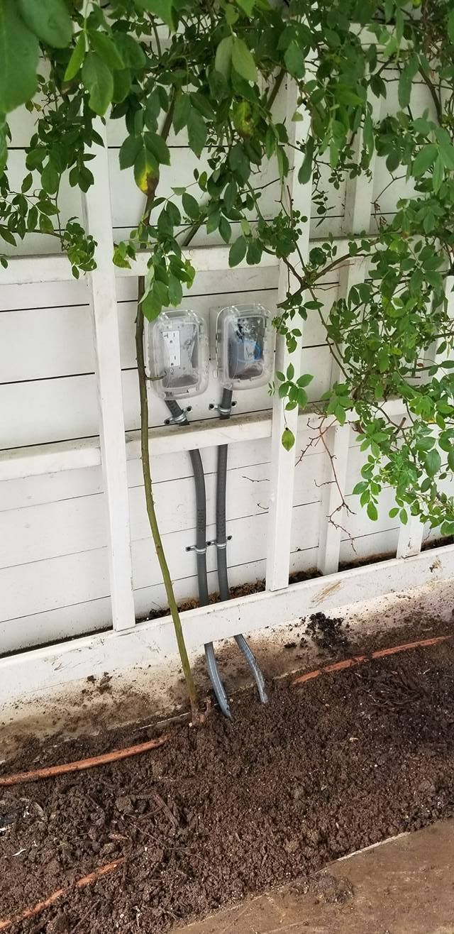 Two electric meters on white wooden fence, with foliage overhead, dark mulch ground.