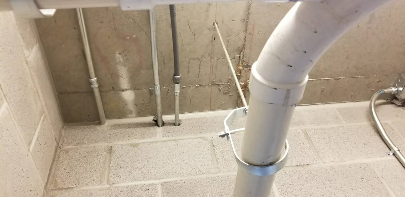 White PVC pipe and metal support bracket against a concrete wall with electrical conduits.
