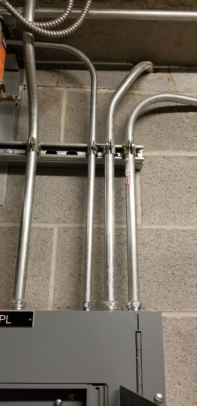 Electrical conduits attached to a gray electrical panel, mounted on a brick wall.