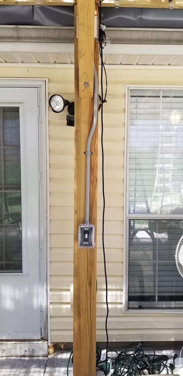 Electrical conduit and outlet box mounted on a wooden post next to a house with a door and window.