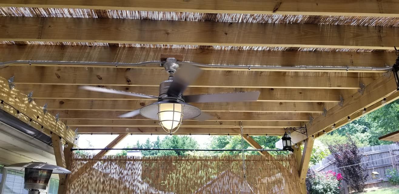 A patio ceiling fan with lights in a wooden pergola, with a thatched roof and plants visible in the background.