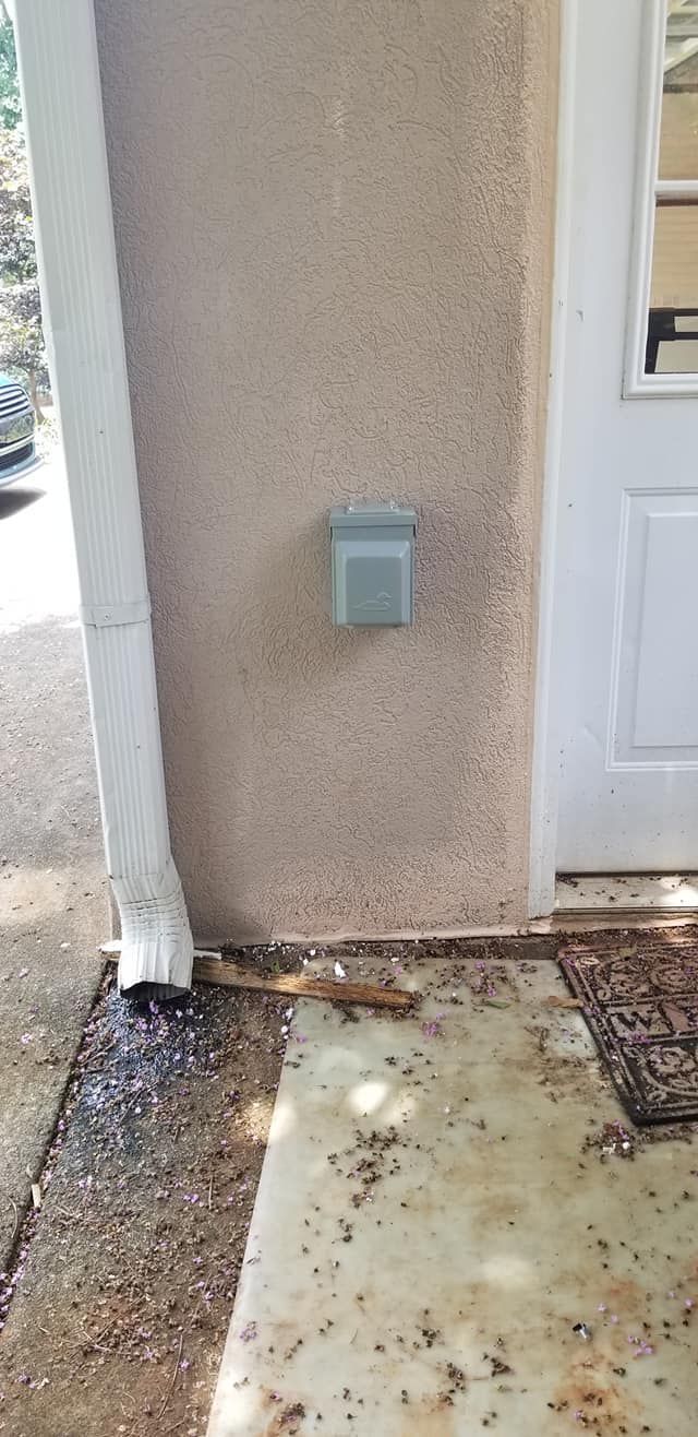A stucco wall with an outdoor electrical outlet, a downspout, and a door. The ground is dirty.