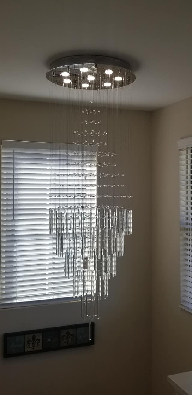 Modern chandelier with cascading crystal prisms hanging near a window with blinds.