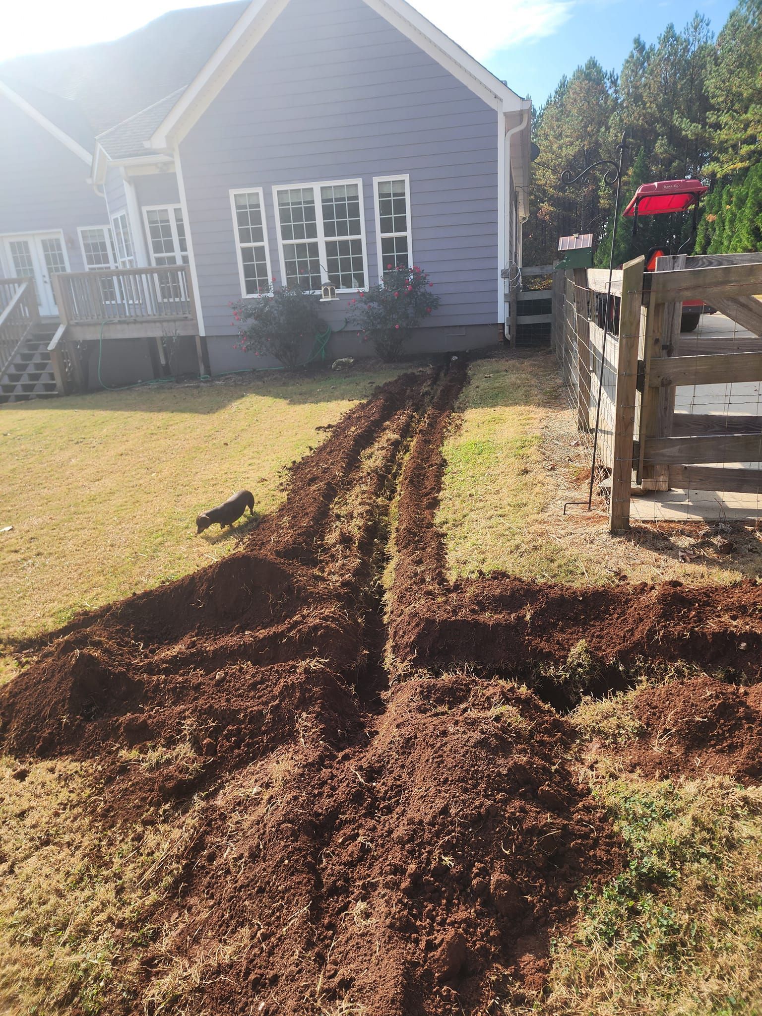 A trench dug in a yard next to a house with a fence and a small deck.