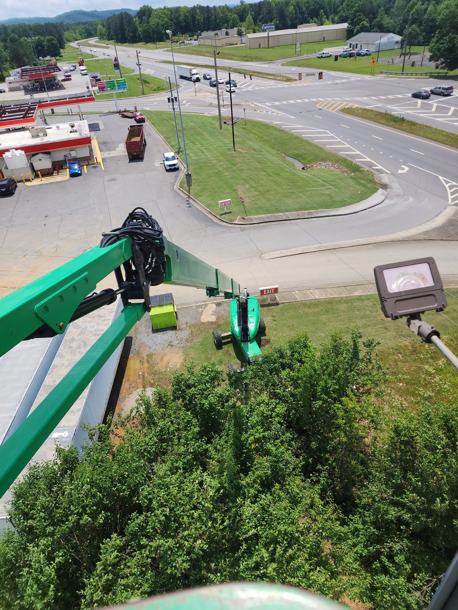 Overhead view of a green boom lift; parking lot, roads, and buildings are in the background on a sunny day.