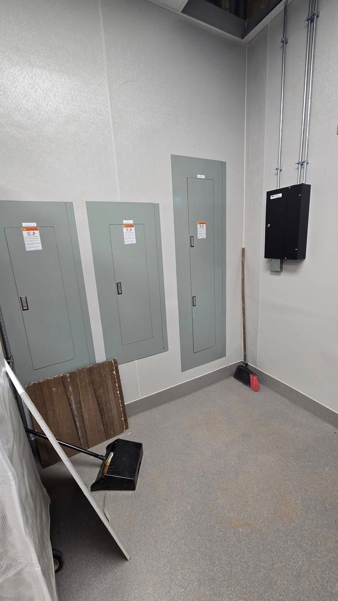 Electrical panels on a wall in a room. Electrical conduit, broom, and equipment present.