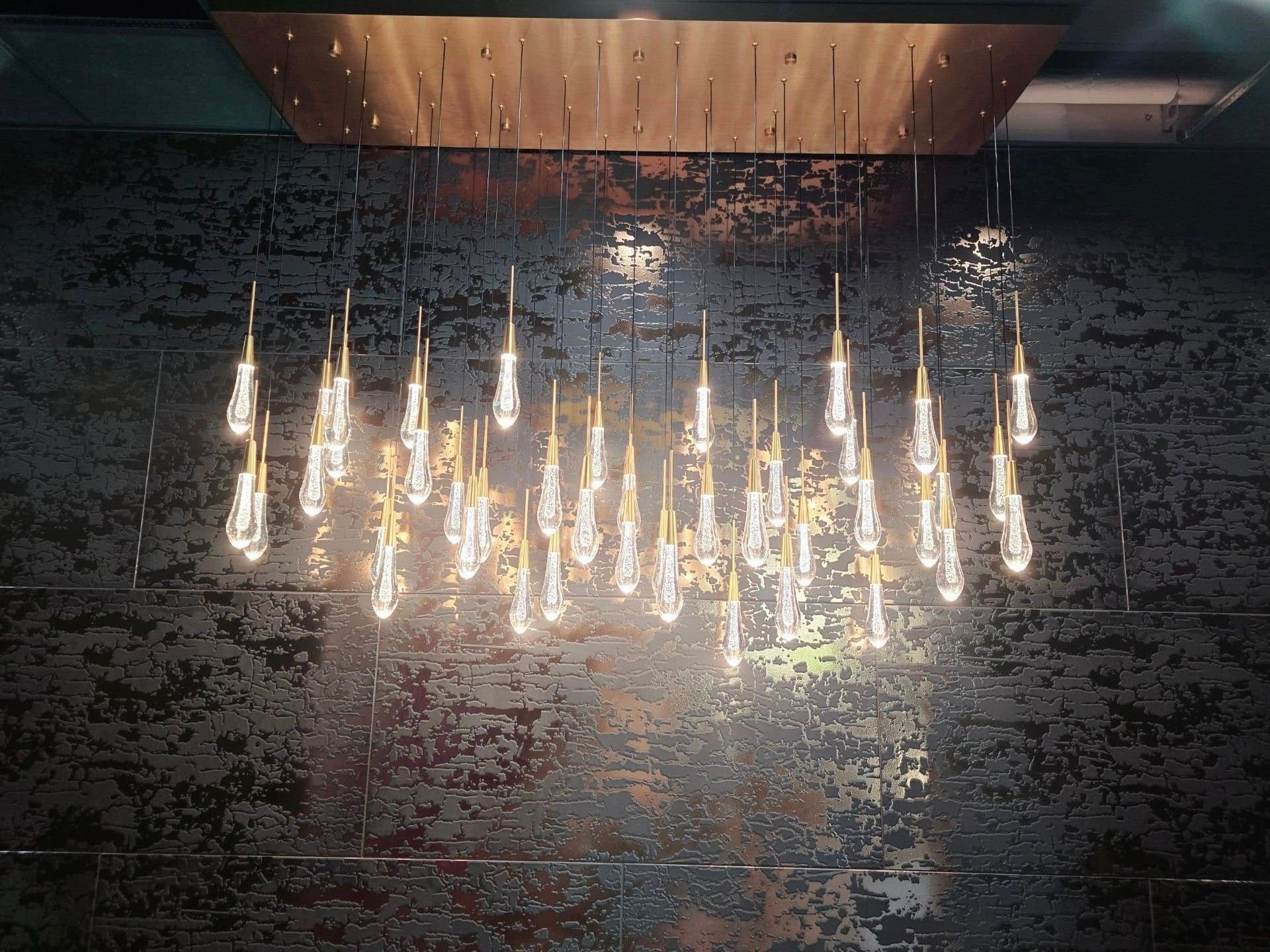 Chandelier with multiple glass teardrop lights hanging from a wooden panel, against a textured dark wall.