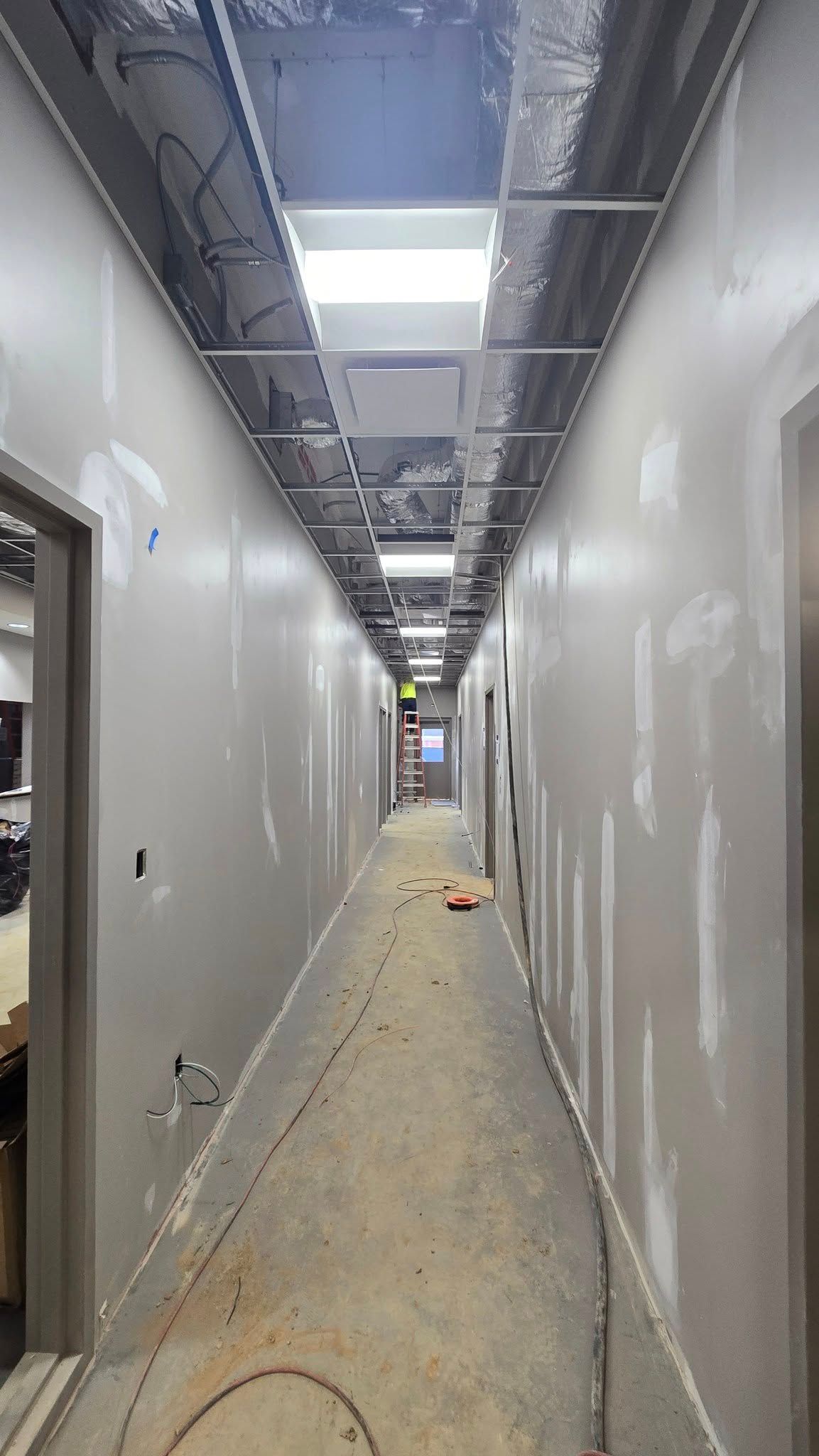 Long, narrow hallway under construction with unfinished walls, doors, and exposed ceiling fixtures.
