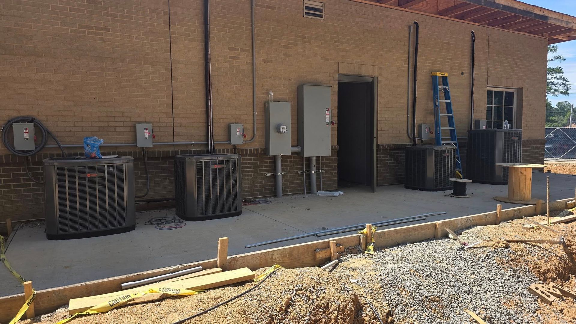 Construction site with air conditioning units and electrical boxes on a brick building exterior. Concrete slab in foreground.