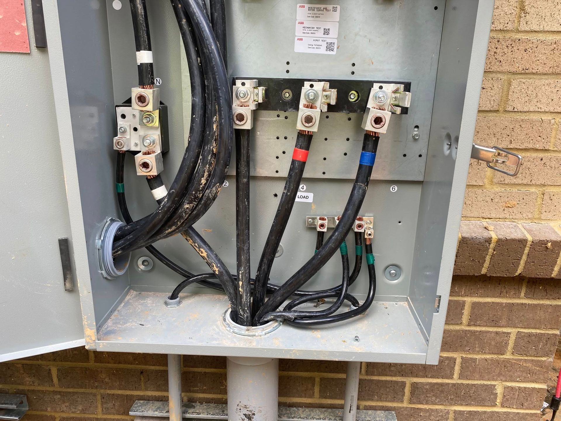 Open electrical panel with three large black cables connected to terminals, near a brick wall.