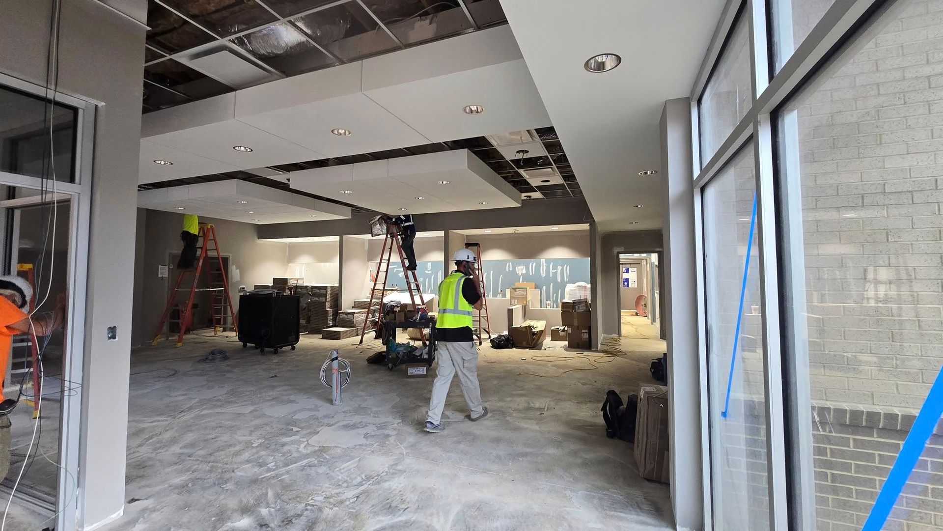 Construction workers inside a building, working on ceiling and lighting. Light concrete floor, partially finished walls.