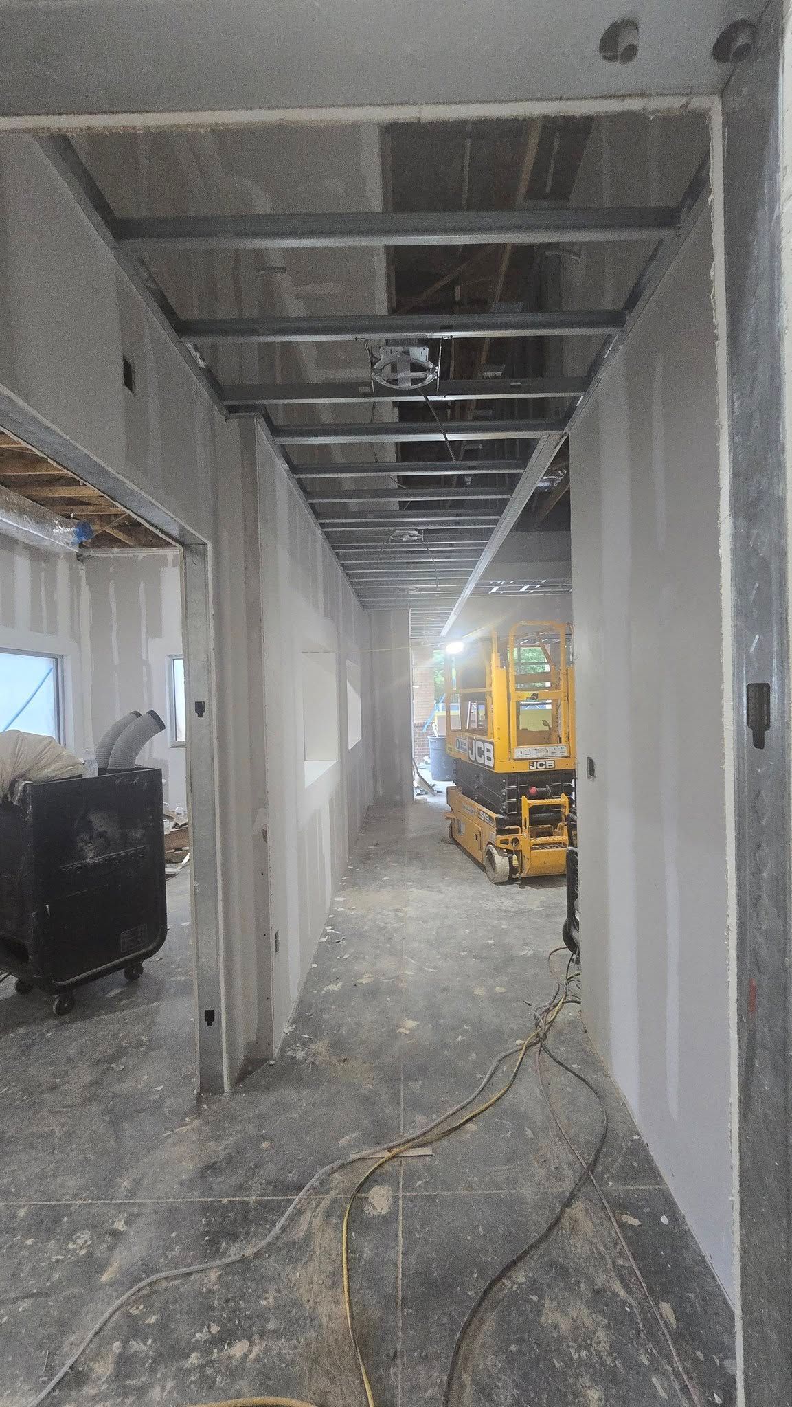 Construction of a narrow hallway with exposed ceiling beams. A yellow lift is visible at the end.