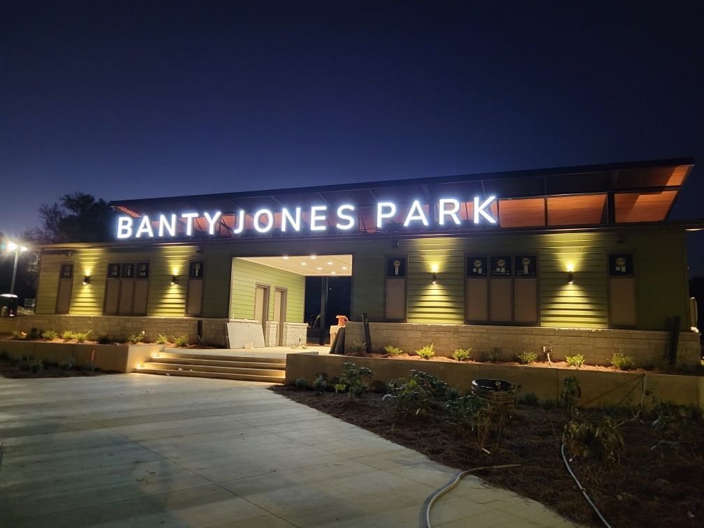 Banty Jones Park entrance at night with illuminated sign. Green building, concrete walkway, and dark blue sky.