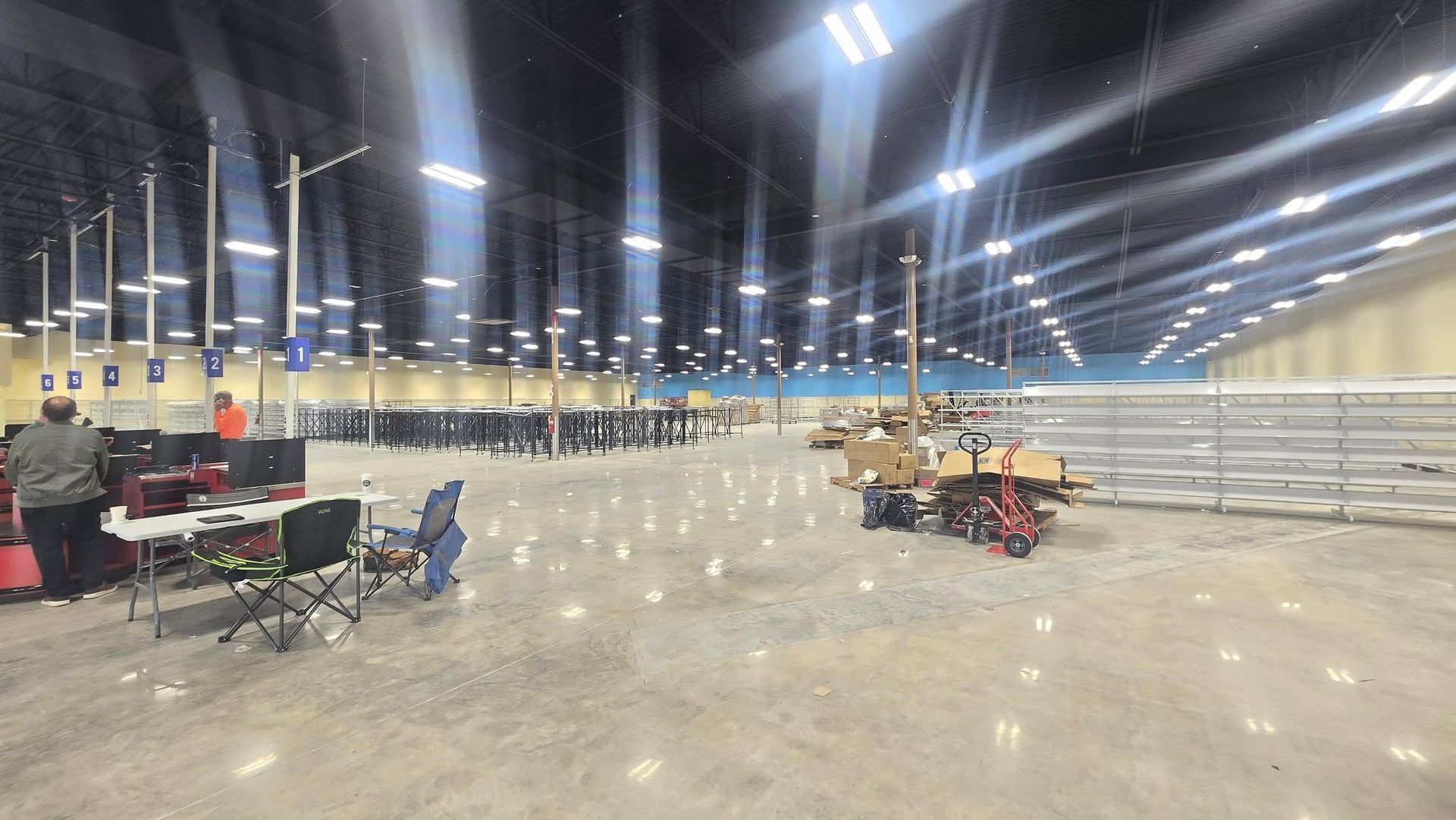 Large, empty warehouse interior with tables, lights, and a few people working.