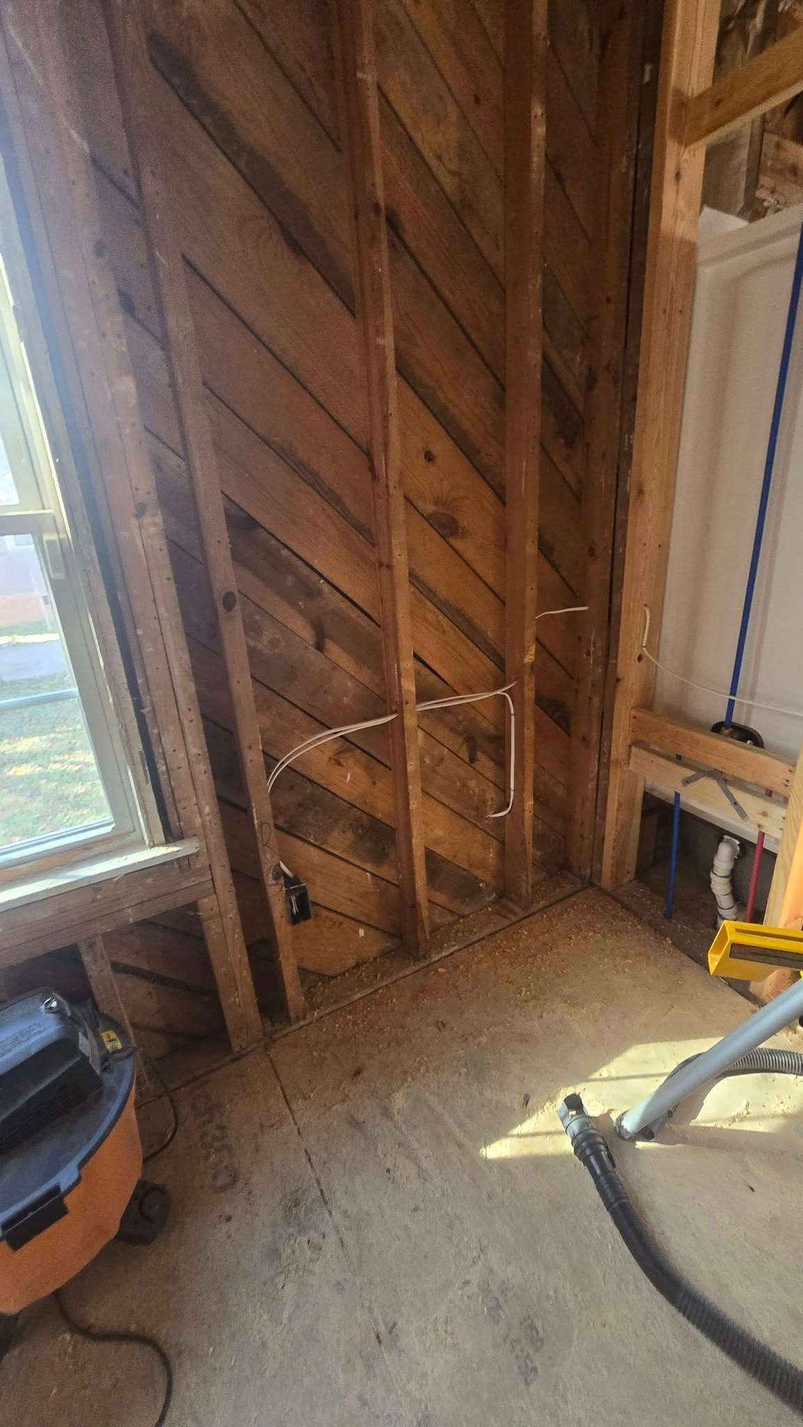 Interior of a building under construction, showing wooden wall studs, exposed pipes, and a window.