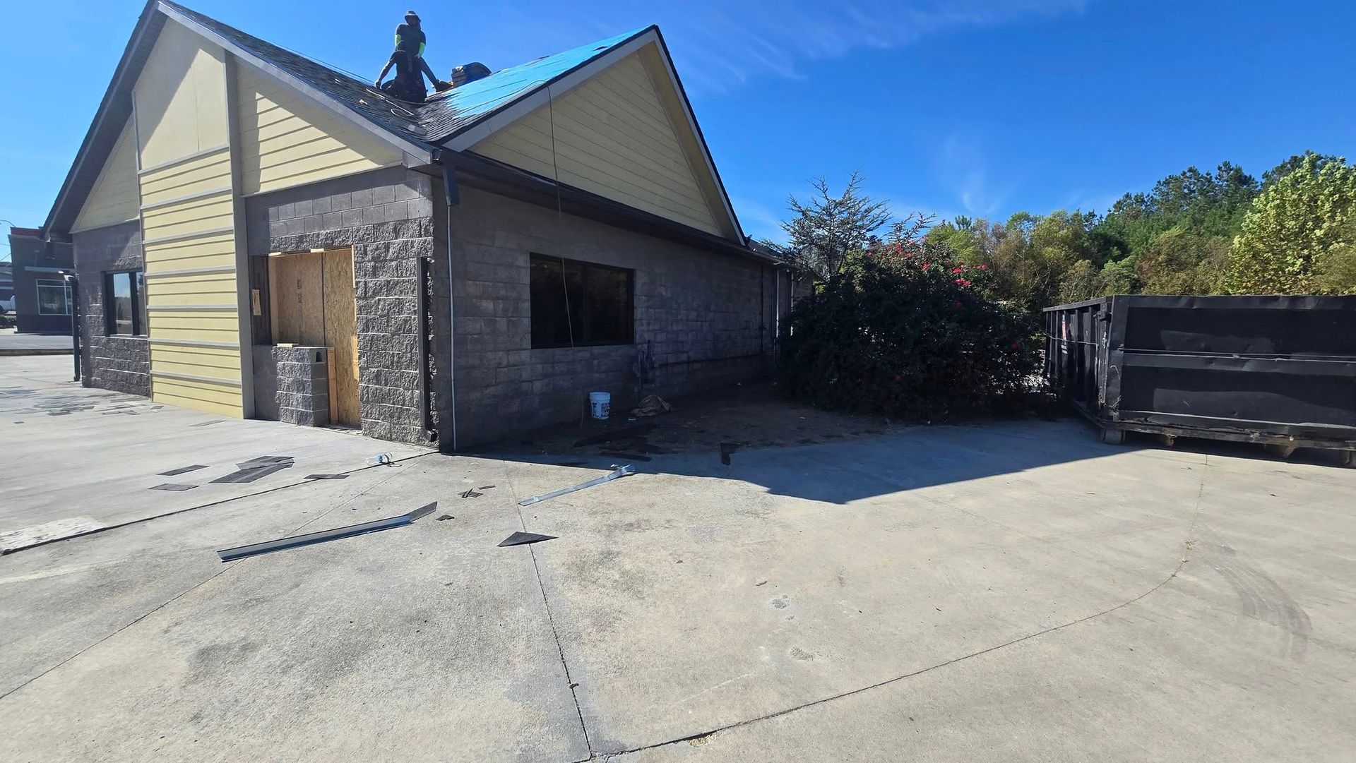 Building undergoing renovation with damaged walls, debris, and dumpster on a concrete lot under a blue sky.