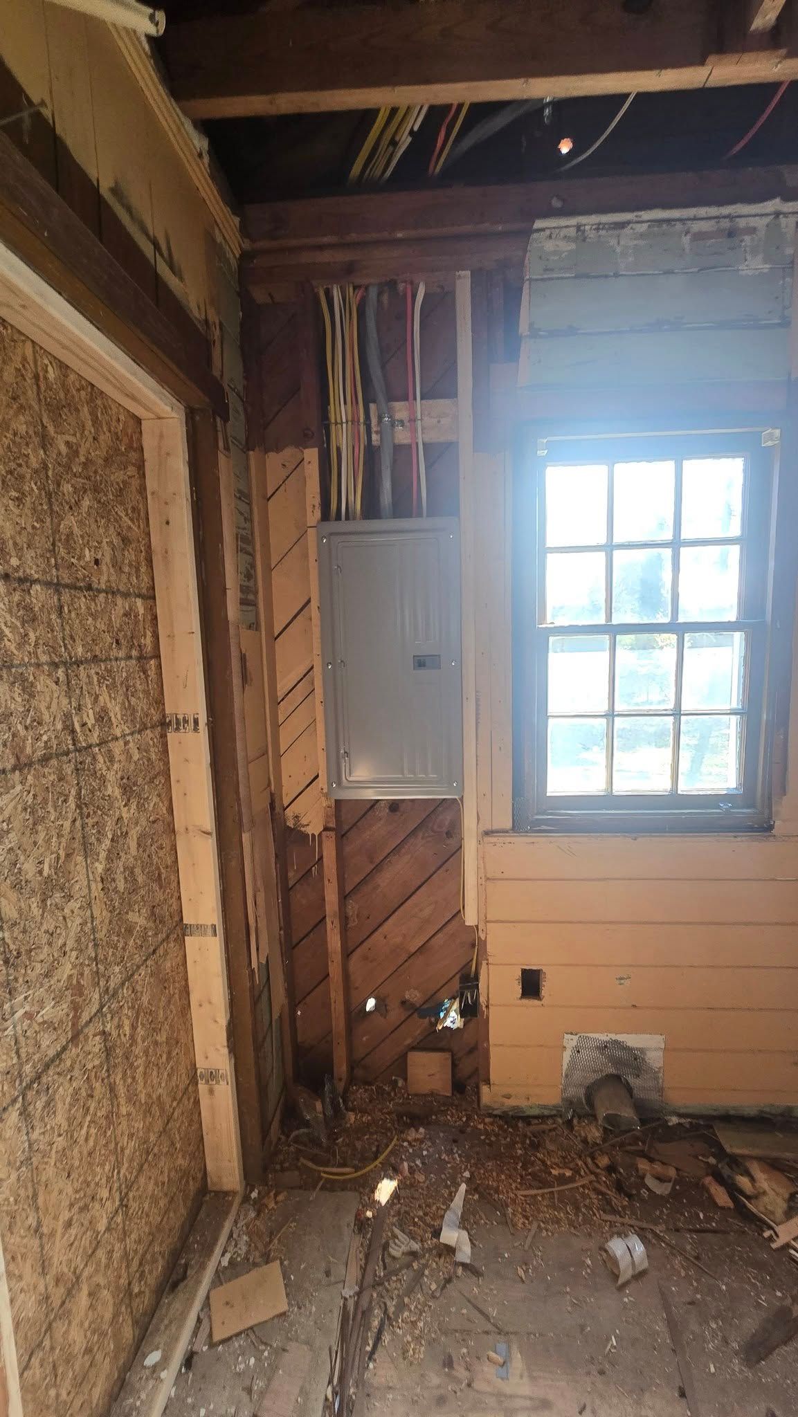 Room interior under construction with exposed walls and electrical panel, window, and debris on the floor.