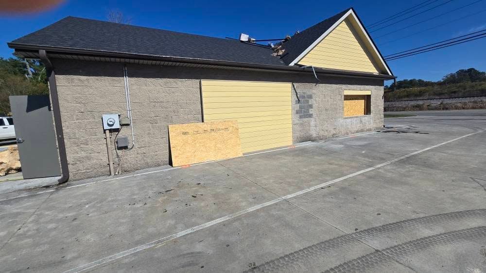 Building with boarded-up windows and garage door. Tan walls, dark roof, light blue sky. Concrete parking area.