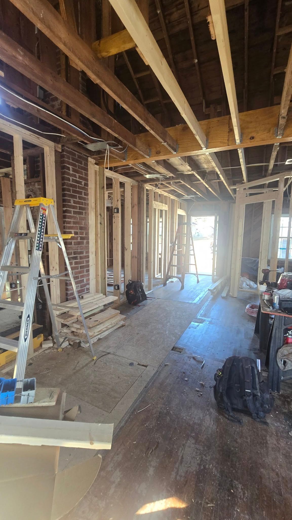 Interior view of a building under renovation, showing exposed framing and brick chimney. Sunlight streams through a doorway.