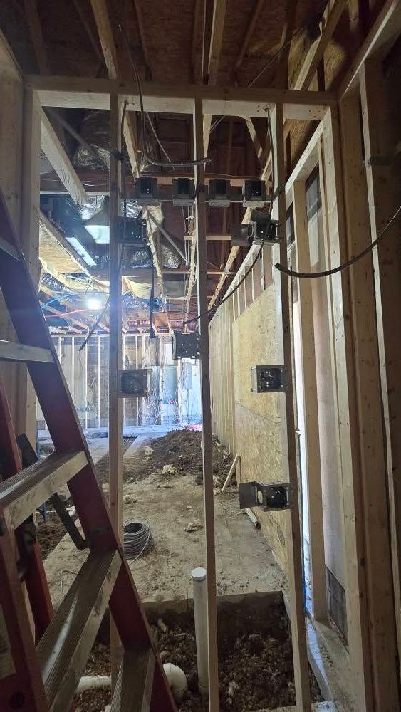 Interior construction site with wooden framing, pipes, and electrical boxes. Ladder leans against the left side.