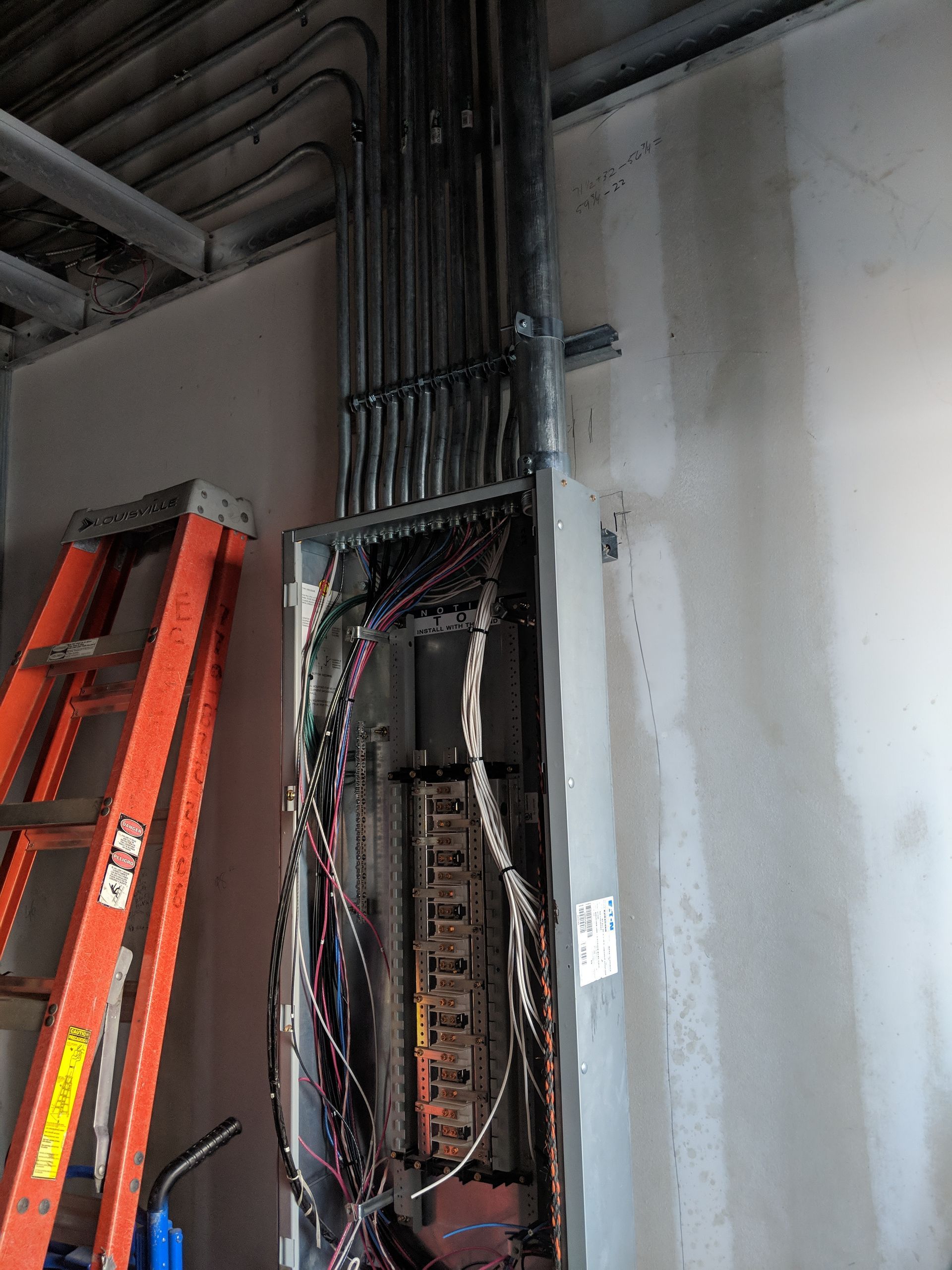 Electrical panel with exposed wires and conduit, adjacent to an orange ladder, mounted on a wall.