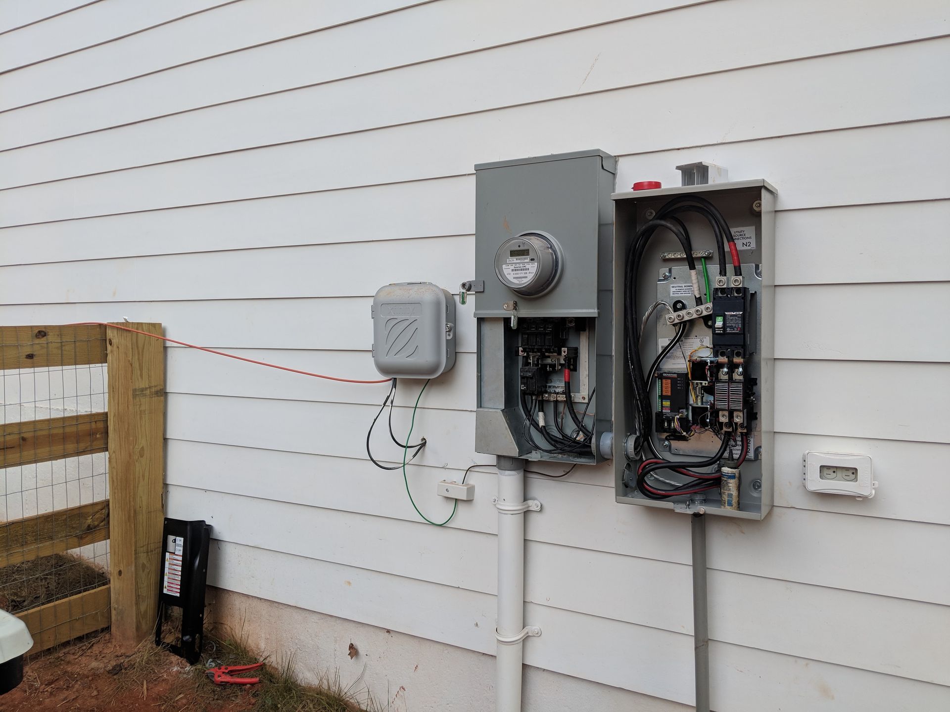 Electrical panel with meter on white siding; open and closed gray boxes with wires.