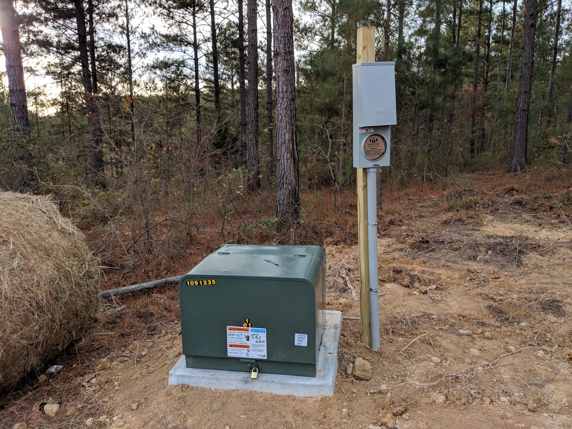 Green electrical box and pole-mounted outlet in a wooded area with a large hay bale.