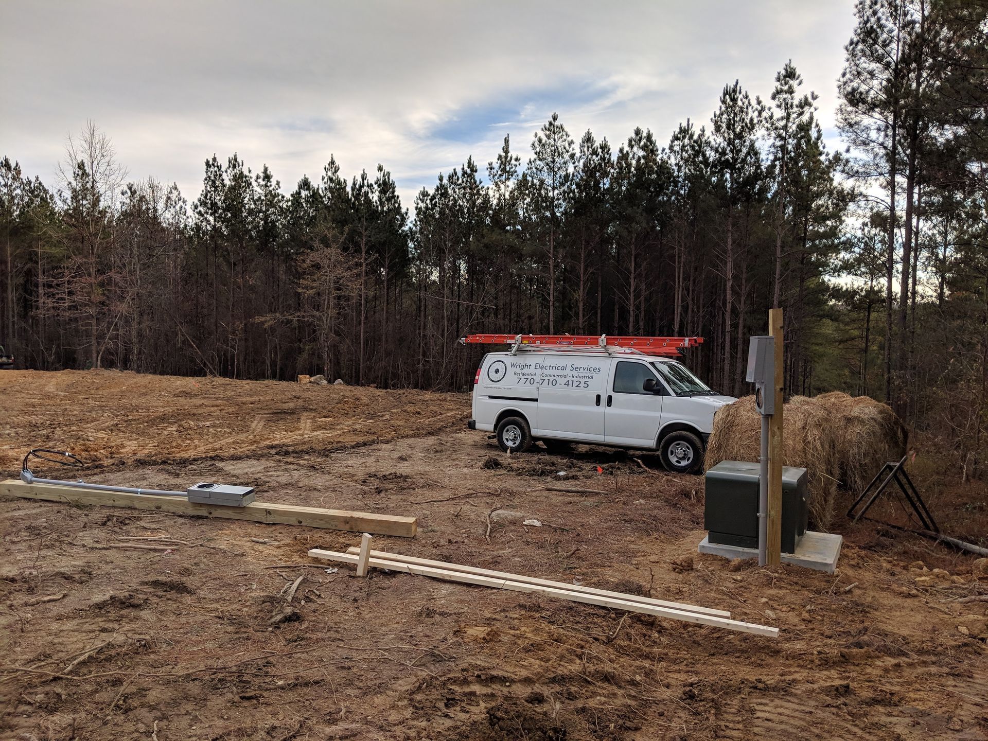White work van parked near a box and forest. Tools and ladder are on the ground.