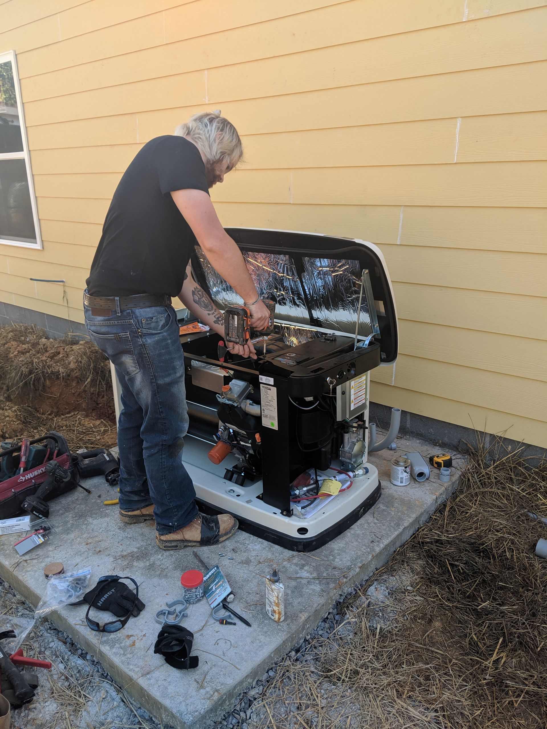 Man working on a generator outside a building. He's wearing jeans and a black shirt. Tools and parts are scattered around.