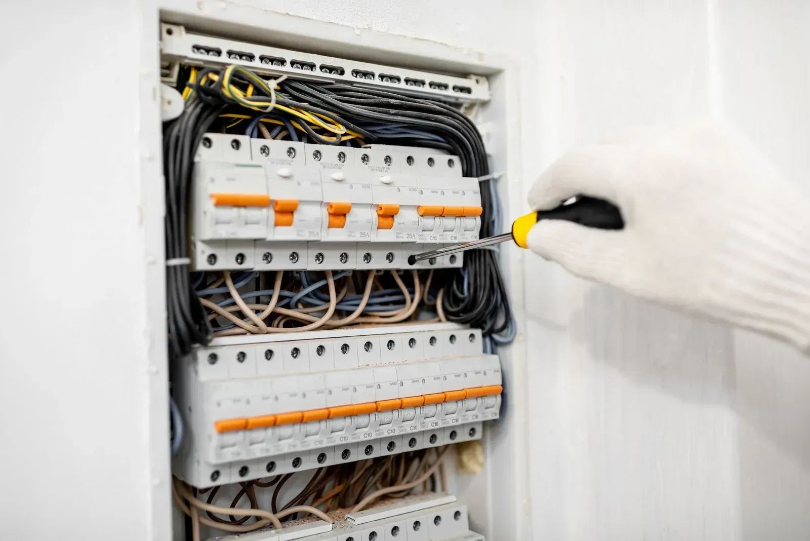 A gloved hand uses a screwdriver on an electrical breaker box with organized wiring and multiple circuit switches.
