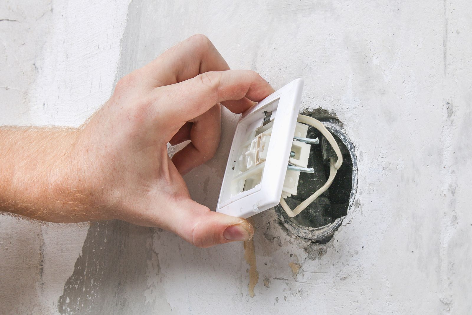 Hand holding a white outlet cover near an exposed electrical outlet in a wall.