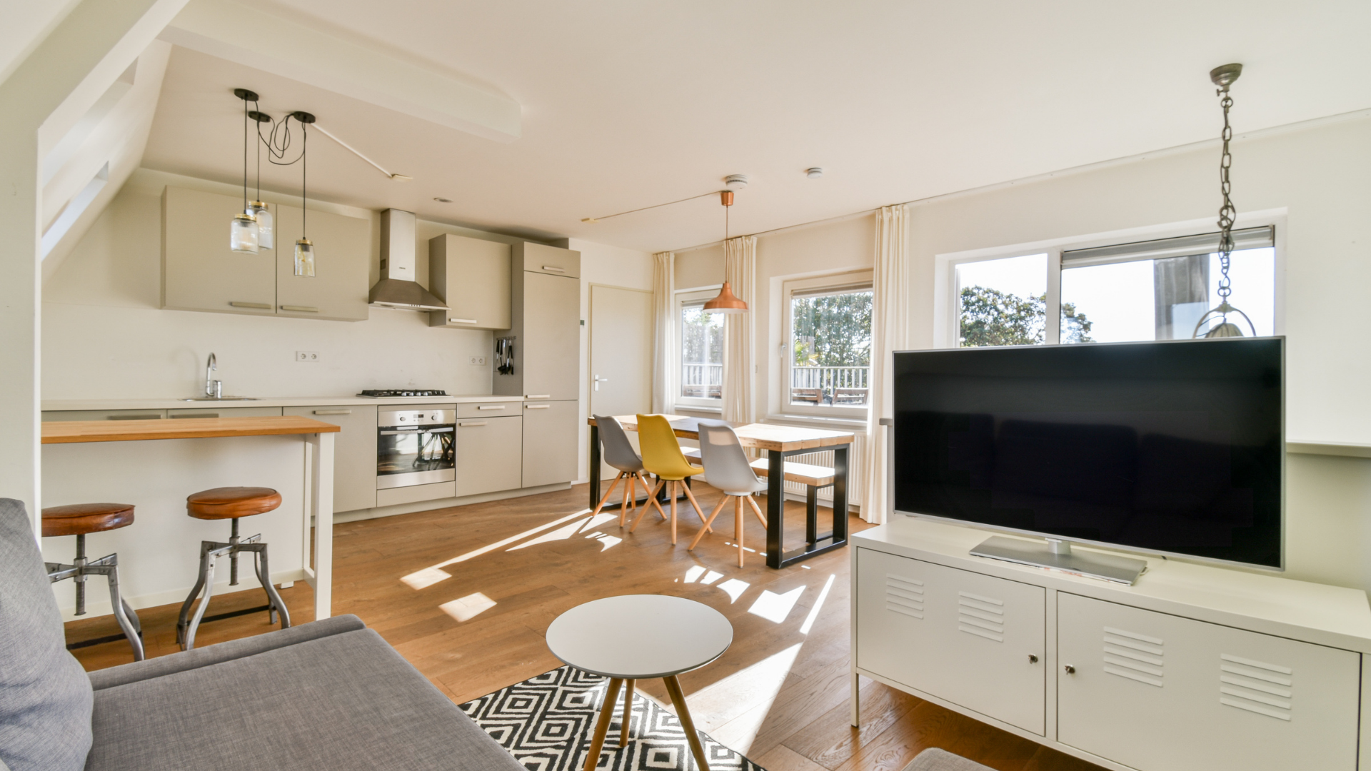 Open-concept living space with kitchen, dining area, and living room. Neutral colors, wooden floors, sunny.