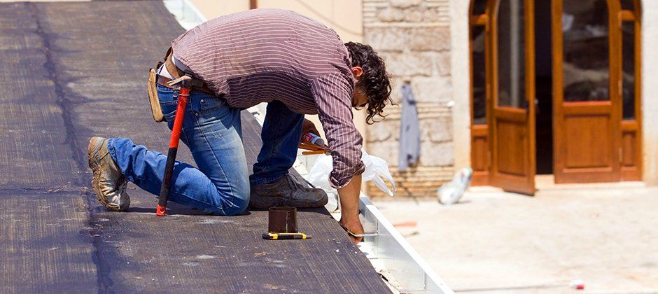 A man is kneeling down on the ground while working on a roof.