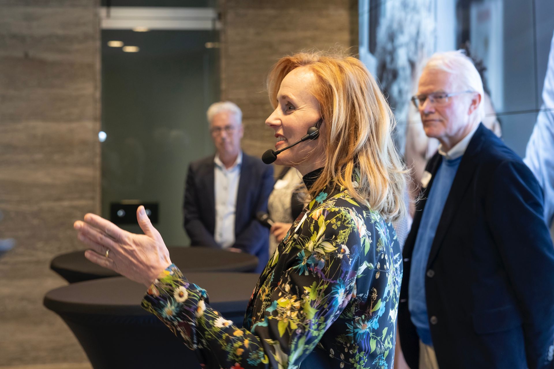 Een vrouw met een microfoon spreekt en gebaart; twee mannen staan achter haar in een modern interieur.