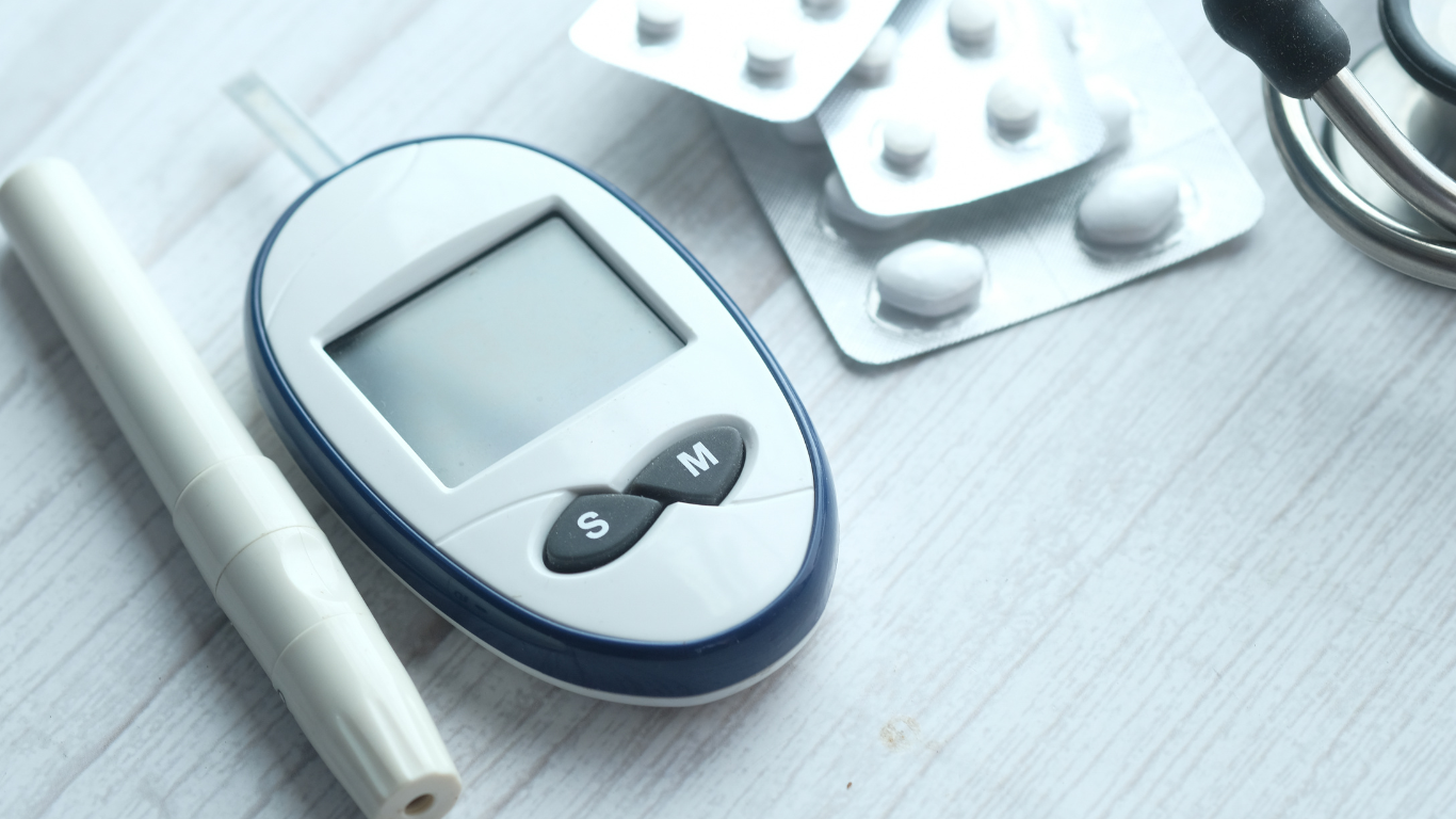A blood glucose meter, a lancing device, and blister packs of white pills rest on a light-colored wooden surface.