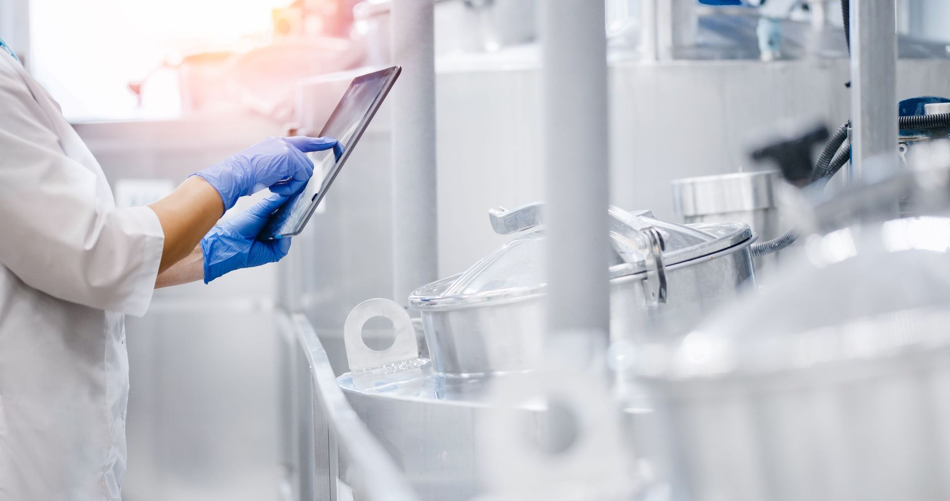 A person wearing blue gloves and a white lab coat uses a tablet in a bright, industrial laboratory setting.