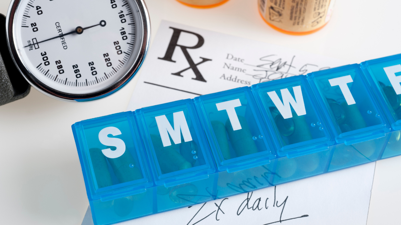 A weekly pill organizer, a blood pressure gauge, and a prescription pad are arranged on a white surface.