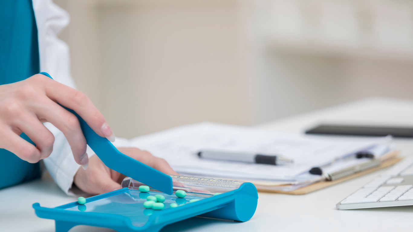 A pharmacist uses a blue spatula to count green tablets on a pill counting tray in a clinical setting.